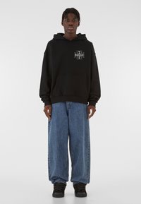 Sort hættetrøje med frontlomme, der har et grafisk design på venstre bryst. Kombineret med løstsiddende blå denimjeans og sorte sneakers.