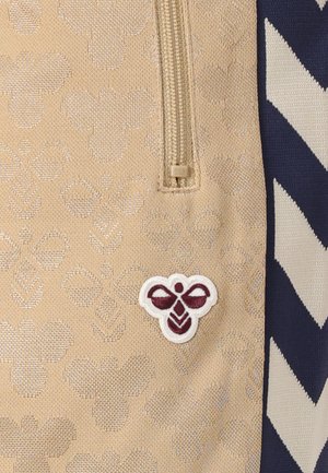 Material bej cu un logo brodat de albină în culoarea marsala și alb, cu un fermoar în partea de sus și un model chevron bleumarin și alb pe partea laterală.