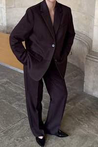Costume taillé marron foncé avec une veste à revers et un pantalon à jambes larges. Des escarpins noirs à bout pointu complètent le look. Tissu lisse, coupe structurée.