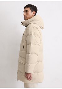 Beige donsjack met afneembare capuchon, doorgestikt ontwerp, zwarte ritsaccenten en een logopatch op de mouw. Zachte textuur en lichtgewicht.