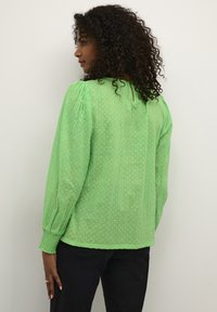Blouse verte texturée avec des manches ballons, arborant un motif à pois et un détail de dos en forme de clé, associée à un pantalon noir.
