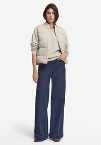 Beige vadderad bomberjacka med hög krage, kombinerad med en ljusgrå tröja och vida mörk denimjeans med uppvikt mudd.