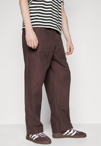 Pantalons amples, marron, en coton, avec des poches latérales et une coupe droite, associés à des baskets violettes et une chemise rayée.