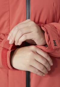Červená zateplená bunda s černým zipem a nastavitelnými manžetami na suchý zip, která vyniká hladkým, texturovaným povrchem.