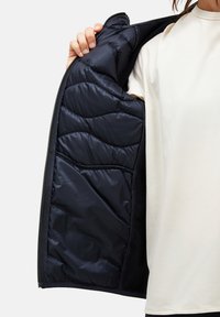 Svart isolerad jacka med vadderad innervadding, som visar en slät yta och strömlinjeformad design. Dragkedjestängning fram.
