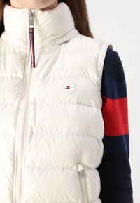 Gilet rembourré léger avec col haut, fermeture éclair, poches latérales et patch logo. Porté par-dessus un pull rayé rouge et bleu marine.
