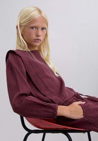 Conjunto texturizado en color marrón oscuro con un patrón de acanalado, que presenta un diseño de hombro drapeado. La modelo está sentada en una silla roja frente a un fondo neutro.