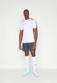 Sportlane seisab, kandmas valget spordisärki mustade triipudega, tumedaid lühikesi pükse, valgeid põlvikuid ning helesiniseid krampe ühtlasel taustal.