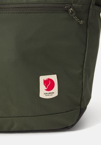 Sac à dos olive avec une texture lisse, équipé d'une fermeture éclair noire et d'un patch de logo de marque rouge et blanc à l'avant.
