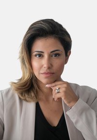 Frau mit braunem, schulterlangem Haar, die einen hellbeigen Blazer �über einem schwarzen Oberteil trägt. Sie hat einen Ring an der linken Hand und berührt ihr Kinn.