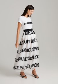 T-shirt blanc avec lettrage noir associé à une longue jupe en tulle à volants comportant des accents de texte noirs. Des talons noirs complètent le look.