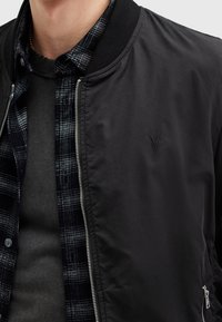 Homme portant une veste bomber noire par-dessus une chemise à carreaux noire et grise et un sous-pull gris foncé, montrant la fermeture éclair et le petit logo de la veste.