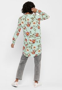 Chemise à manches longues couleur menthe avec un motif floral rose et jaune, associée à un jean gris et des chaussures blanches, présentant un design à ourlet arrondi.