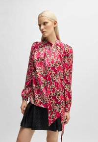 Fuchsia-blumiges Oberteil mit langen Ärmeln, Stehkragen und drapiertem Design. Ein strukturierter schwarzer, ausgestellter Rock ergänzt das Oberteil.