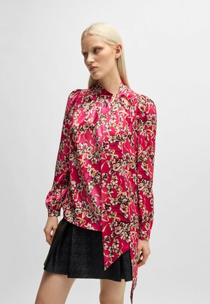 Fuchsia-blumiges Oberteil mit langen Ärmeln, Stehkragen und drapiertem Design. Ein strukturierter schwarzer, ausgestellter Rock ergänzt das Oberteil.