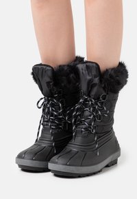 Botas de nieve impermeables negras presentan un upper acolchado con ribete de piel sintética, diseño de cordones y una suela de goma gruesa para tracción.