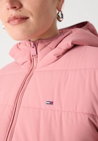 Roze pufferjack met een capuchon, voorzien van horizontale stiksels en een klein merklogo in rood, wit en blauw nabij de rits.
