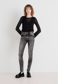 Top sans manches noir avec des manches longues en dentelle, associé à un jean gris taille haute. Le modèle porte des chaussures à platforme noires. Fond minimaliste.