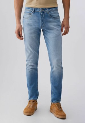 Uomo che indossa jeans aderenti azzurri chiari e sneaker in suede color cammello, in piedi contro uno sfondo chiaro semplice.