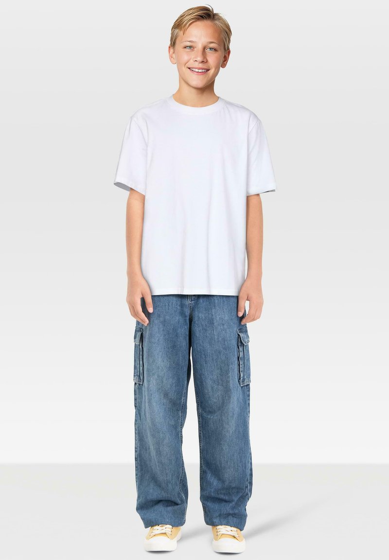 Lächelnder Junge mit kurzem blonden Haar, der ein einfaches weißes T-Shirt, lockere blaue Cargo-Jeans und gelb-weiße Sneaker trägt, vor einem grauen Hintergrund steht.