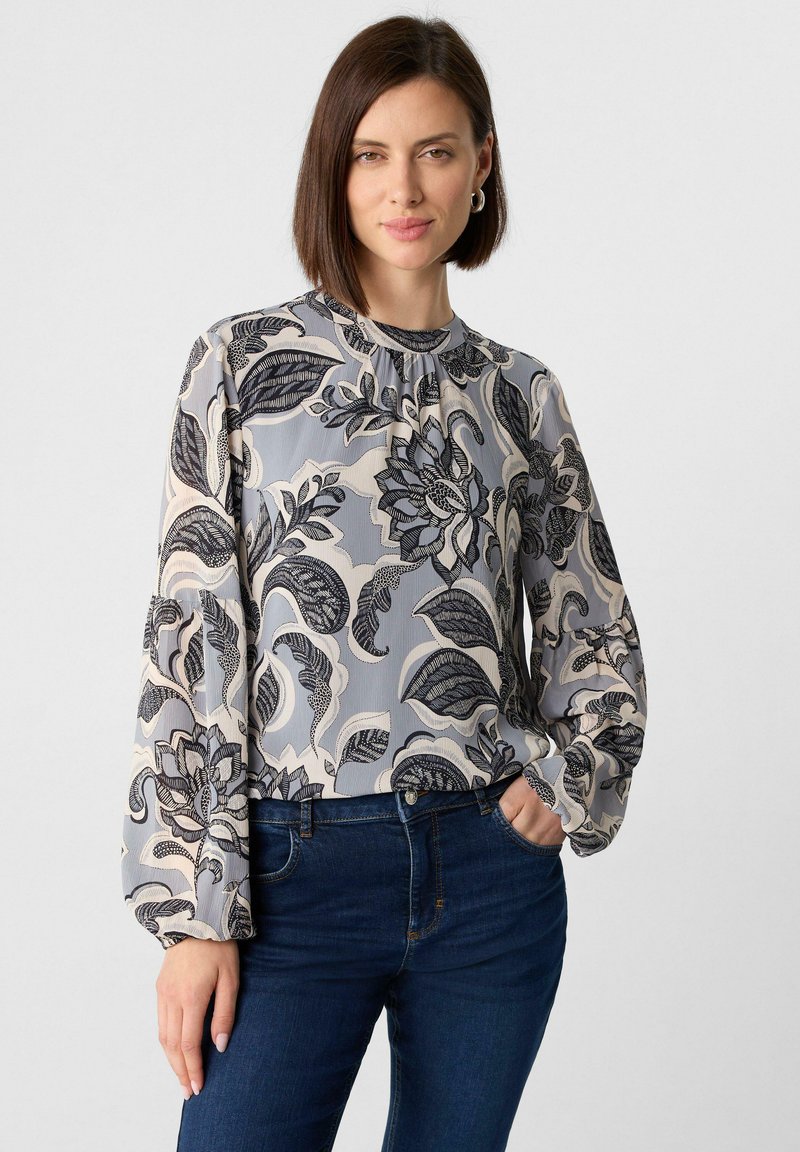 Blumenbluse in Hellgrau mit dunklen Blumenmustern, langen Puffärmeln und einem runden Halsausschnitt. Kombiniert mit dunkelblauen Jeans.