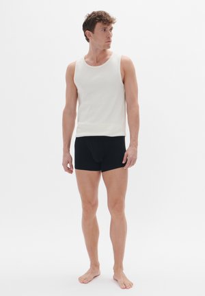 Witte geribde tanktop met een ronde hals, gecombineerd met zwarte strakke shorts. Het model staat blootsvoets tegen een effen achtergrond.