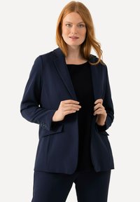 Blazer sartorial blu navy con rever a punta, due tasche frontali e polsini con tre bottoni. Tessuto liscio con una vestibilità strutturata.