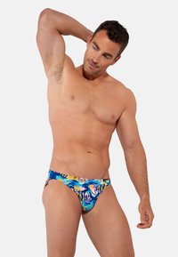 Muskelmann posiert mit der Hand hinter dem Kopf, trägt bunte Schwimmbrieftaschen mit abstraktem Muster vor einem schlichten hellen Hintergrund.