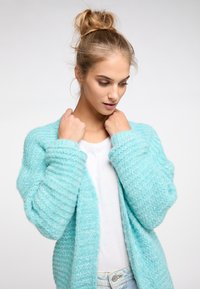 Hellblauer, strukturierter Cardigan mit lockerer Passform und weiten Ärmeln. Getragen über einem weißen Hemd, das einen weichen, kuscheligen Stoff zur Schau stellt.