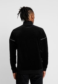 Schwarze Veloursjacke mit hohem Kragen, ausgestattet mit weißem Piping entlang der Schultern sowie gerippten Bündchen und Saum. Glatte Textur und taillierte Passform.