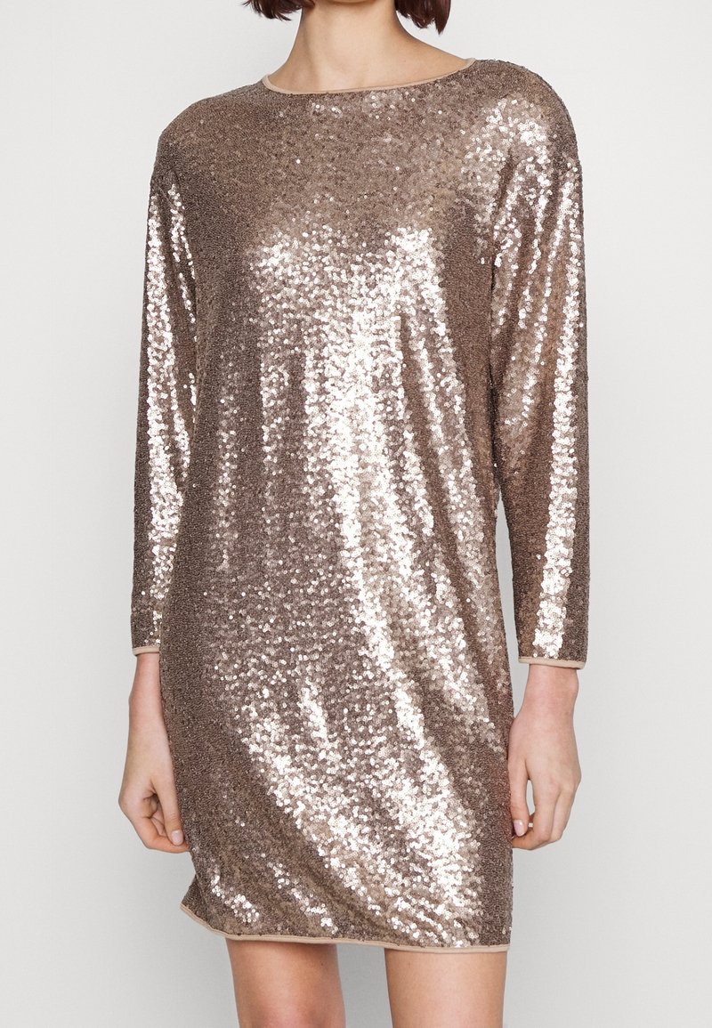 Femme portant une robe à paillettes rose doré brillante, longueur genoux, manches longues, col rond, debout devant un fond clair uni.