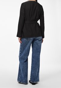 Blazer noir ceinturé à manches longues, associé à un jean bleu clair à jambes larges avec une texture délavée et un ourlet effiloché. Chaussures noires visibles.