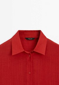 Chemise en lin rouge dotée d'un col classique, de deux boutons et d'une surface texturée, adaptée pour une tenue décontractée ou formelle.