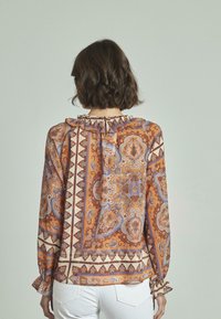 Blusa a maniche lunghe arancione con motivo paisley, caratterizzata da un colletto e polsini arricciati, realizzata in tessuto leggero. Vista da dietro.