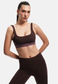 Brun sport-bh med en lavendelfärgad kant, med fyrkantig halsringning och breda axelband, i kombination med högmidjade bruna leggings.