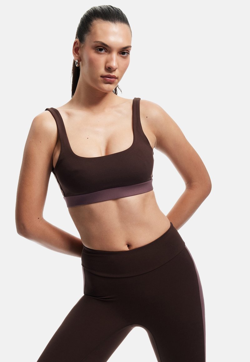 Sujetador deportivo marrón con una banda lavanda, con escote cuadrado y tirantes anchos, combinado con leggings marrones de cintura alta.