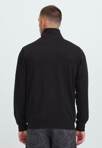 Mann med kort hår og hoop-øredobber iført en svart høyhalset genser og falmede svarte jeans, sett bakfra mot en ensfarget bakgrunn.