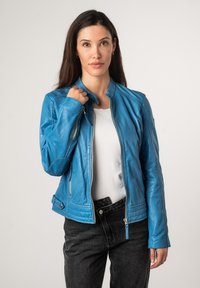 Blaue Lederjacke mit Reißverschluss, taillierter Schnitt, Seitentaschen und gerippten Akzenten an den Schultern. Getragen über einem weißen Hemd und schwarzen Jeans.