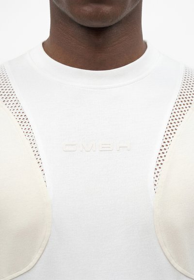 Κοντινό πλάνο ενός ατόμου που φοράει λευκό μπλουζάκι με ανάγλυφο κείμενο "GMBH", με μπεζ επενδυμένες πλέγμα ενθέσεις στους ώμους.