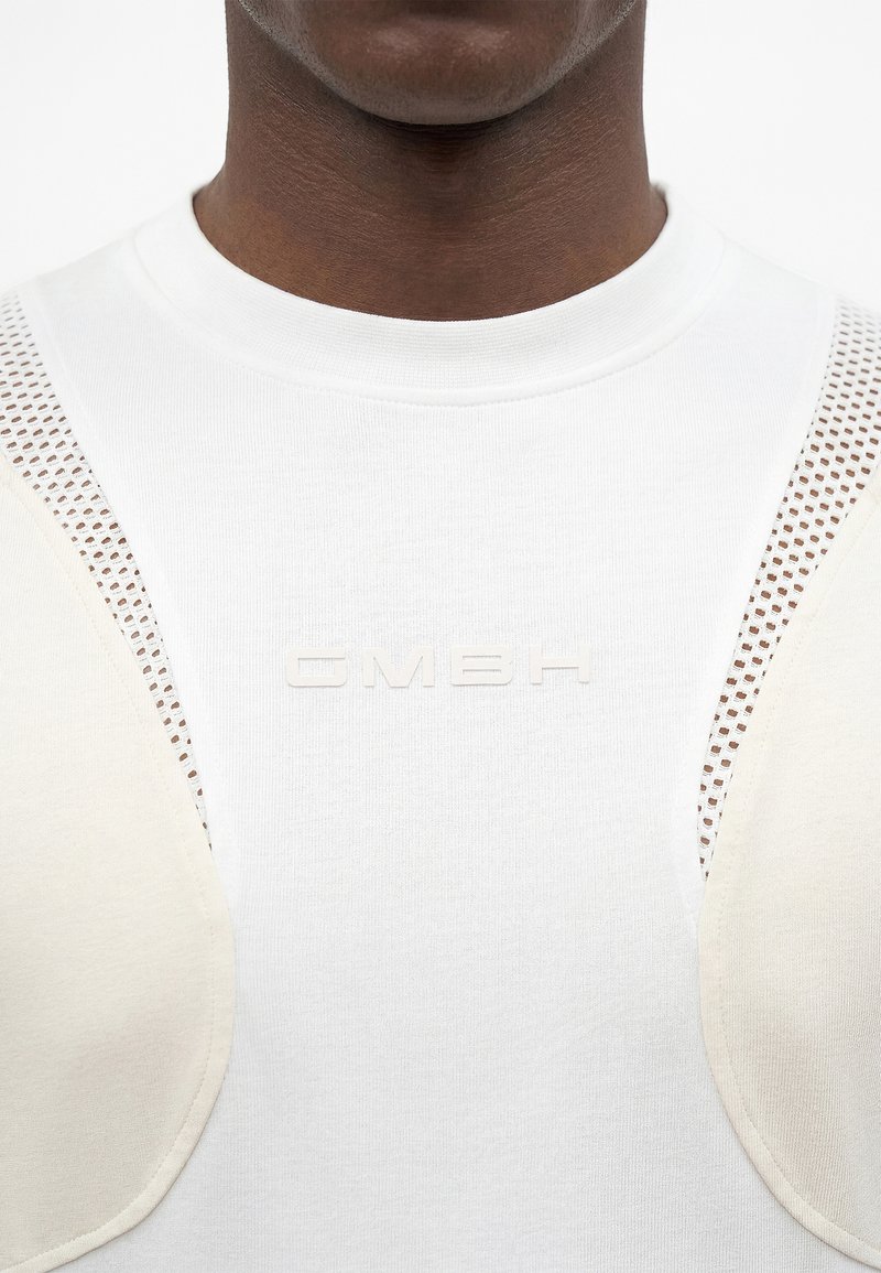 Gros plan d'une personne portant un t-shirt blanc avec le texte en relief "GMBH", doté d'inserts en mesh beige rembourrés sur les épaules.