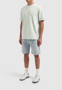 Lichtgroen katoenen T-shirt met korte mouwen en een ronde hals, gecombineerd met lichtblauwe denimshorts. Witte sneakers met grijze accenten.