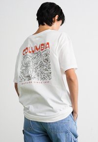 Person with short dark hair wearing a white Columbia T-shirt featuring a mountain design and text "Outside since 1938," paired with blue pants.