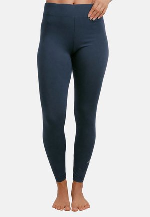 Kvinde iført marineblå høj-taljede leggings, stående barfodet, med den ene hånd hvilende på hoften, mod en ensartet lys baggrund.