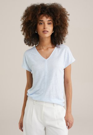 Helles blaues T-Shirt mit kurzen Ärmeln und V-Ausschnitt aus weichem Stoff, mit einer dezenten Textur und lockerer Passform. Kombiniert mit weißen Hosen.