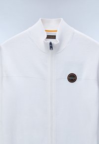 Vit zip-up jacka med en texturerad övre panel och slät undre sektion, med en rund logobadge och ståkrage.