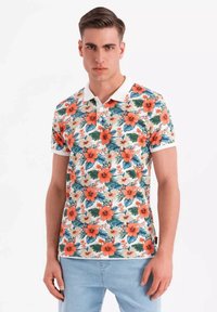Blommig polo-skorta med orange hibiskus och gröna blad på en krämfärgad bakgrund, med en vit krage och korta ärmar.