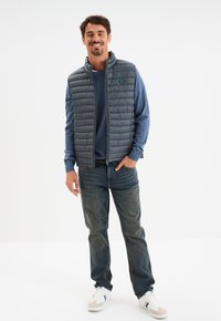 Graue Steppweste mit horizontaler Steppung, Stehkragen, über einem blauen Langarmshirt. Slim Fit Jeans und weiße Sneakers runden das Outfit ab.