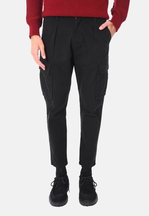Pantaloni cargo neri con pieghe frontali, due tasche applicate sui lati, vestibilità affusolata, realizzati in tessuto resistente, abbinati a scarpe nere e a un maglione rosso.
