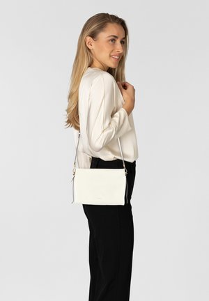 Femme aux longs cheveux blonds portant un chemisier blanc et un pantalon noir, avec un sac bandoulière blanc sur l'épaule, souriant de profil.