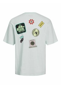Camiseta de manga corta verde menta con varios parches coloridos que incluyen texto y diseños, con un tema de hotel y signos de dólar.